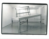 A sink structure on wheels sits atop a tilted wooden table top with legs of varying sizes. Light from the ceiling is seen emanating onto the structure in the white-walled gallery space.