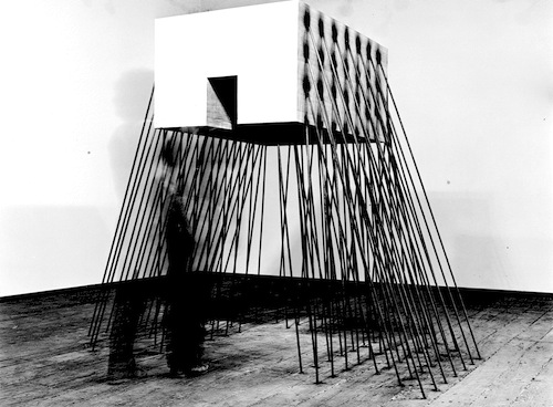 A large square edifice with an opening in its center bottom held up off the floor with numerous steel rods on its sides in a gallery setting. A shadow of a human figure appears/disappears in front of the work.