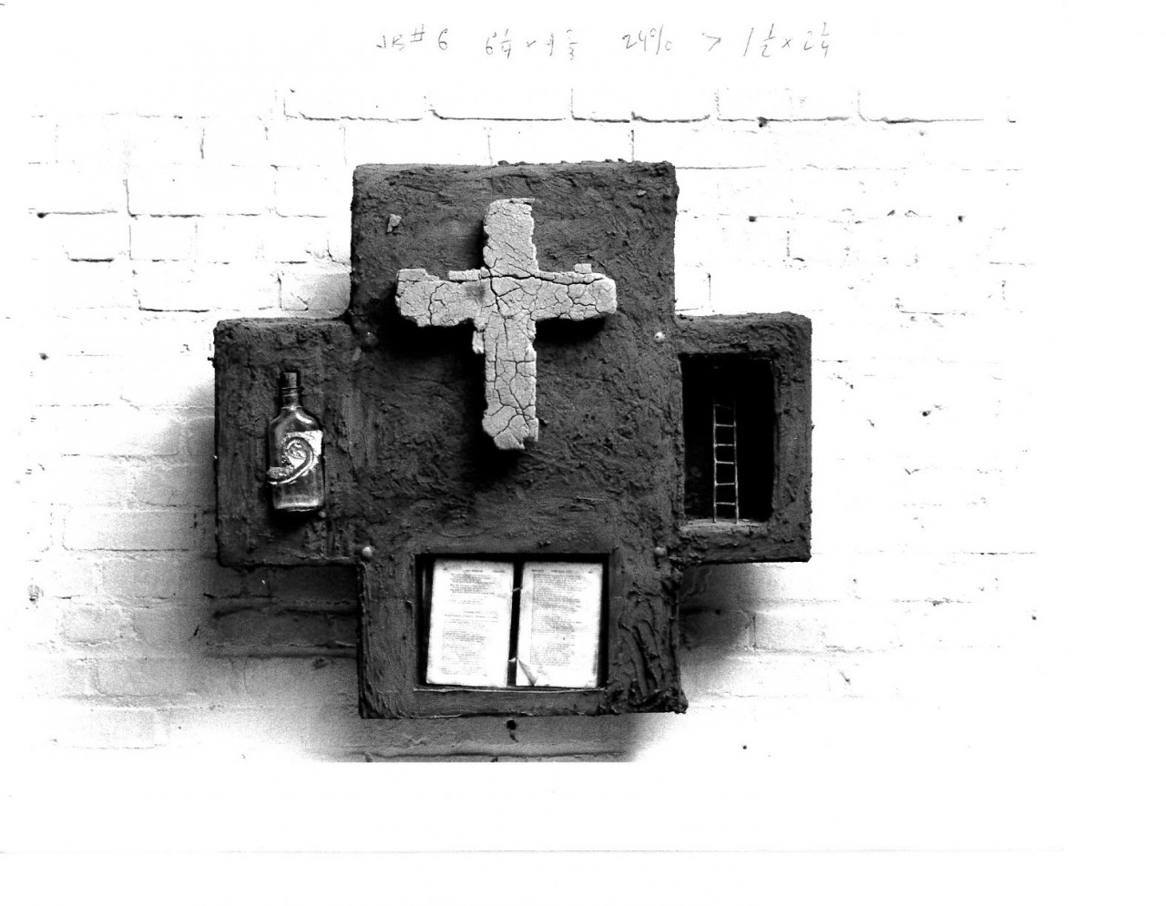 A sculpted cross appears on a larger sculpted cross along with a small bottle on its left, a ladder inset on the right and an open text on the bottom part. All is hung on a white gallery wall and dimensions are written above the sculpture in pencil.
