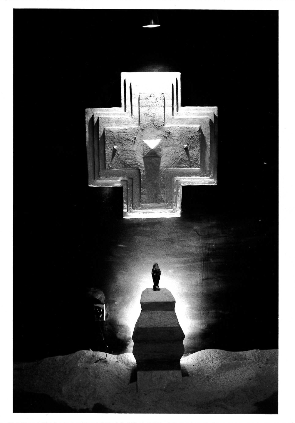 A sculpted pedestal with a black object on it is visible below a sculpted multilayered cross hanging on a gallery wall. Sharp lighting shrouds most of the image in darkness with a light falling on the figure on the pedestal.