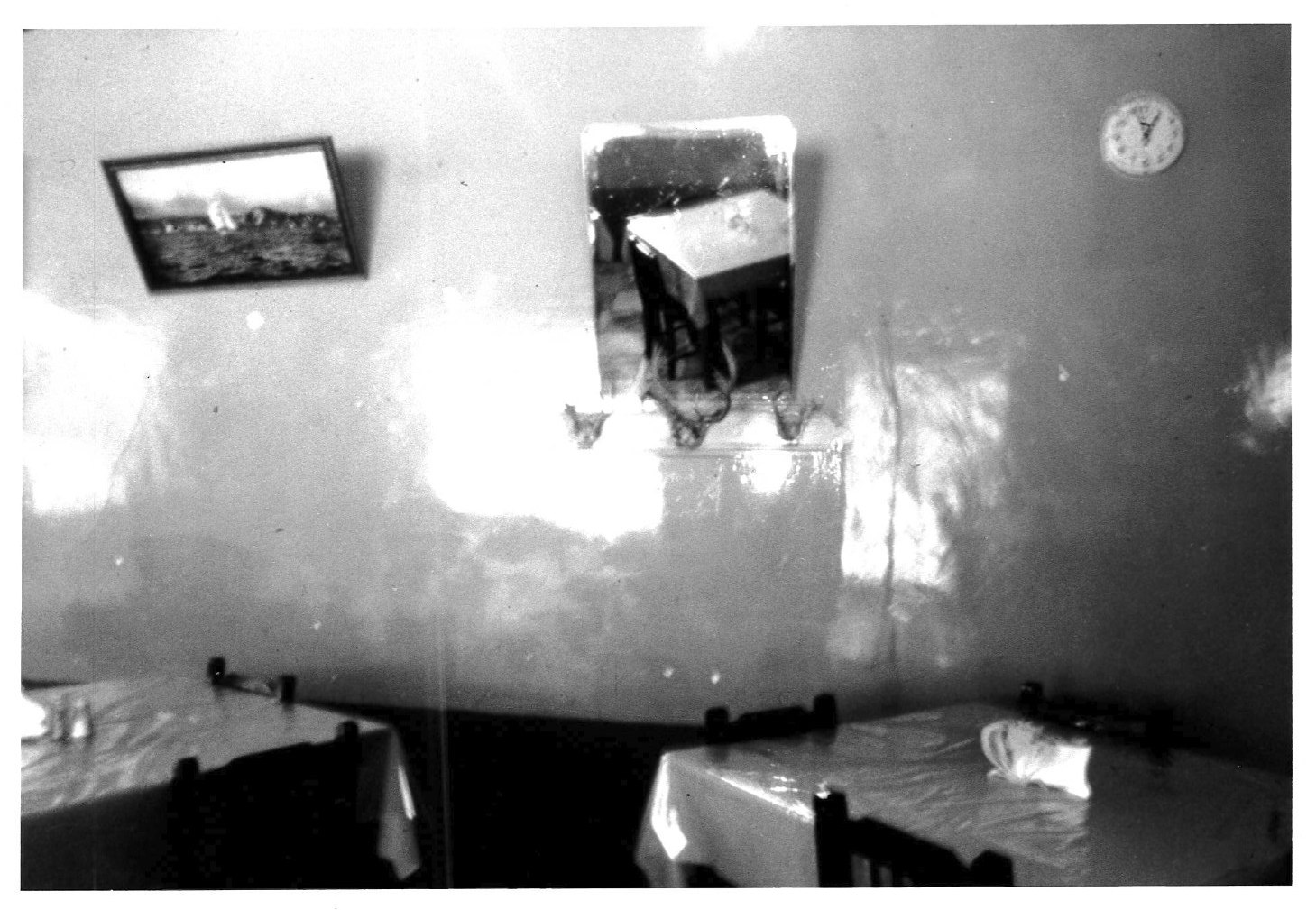 Two beds appear on the floor with a mirror and a picture hanging on the walls along with a clock in the right corner. Light is shining onto the wall from an area not visible to the viewer.