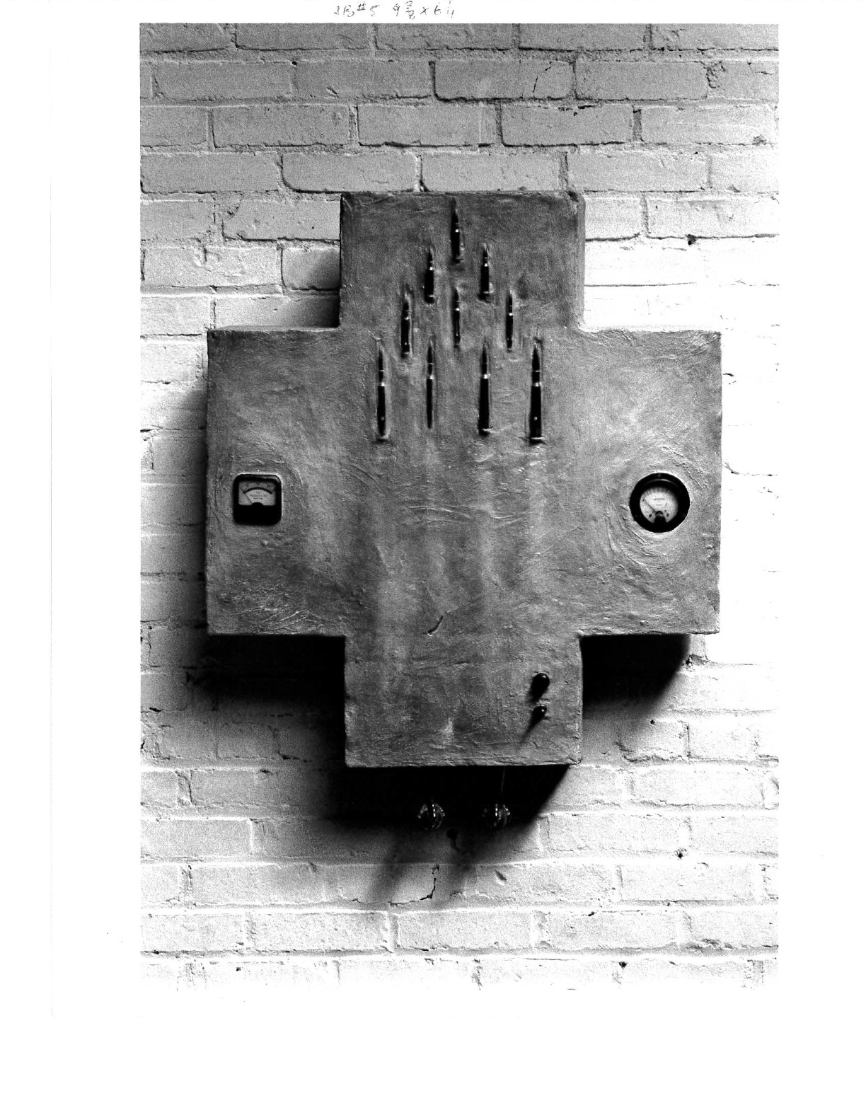 A sculpted cross with two scale readings on either side of it along with bullets lined in formation on the top half and some undefinited black marks on the bottom half and two unidentified objects hanging underneath. It hangs on a white brick wall.