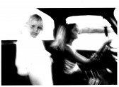 A child's face with a blurred white body stands next to a figure driving a car with one hand on the wheel and another outside of the window. The outside of the car is also white in appearance.