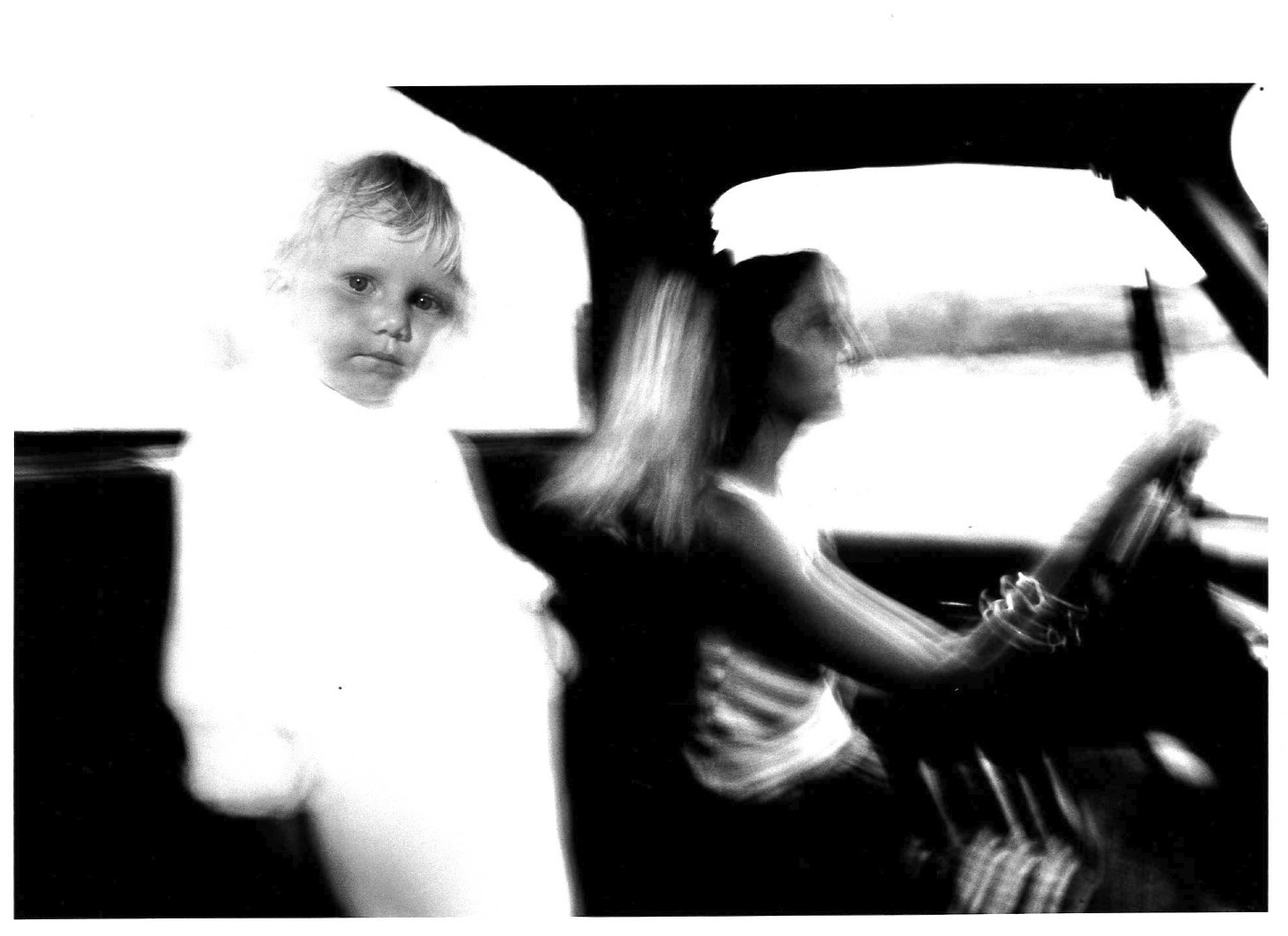 A child's face with a blurred white body stands next to a figure driving a car with one hand on the wheel and another outside of the window. The outside of the car is also white in appearance.