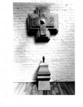 A sculpted pedestal with a black object on it is visible on a wooden floor below a sculpted multilayered cross hanging on a white gallery wall. Numbers are visible above the image.