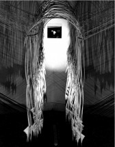 A spotlight shines on a photograph hanging on a white wall framed by string and electrical wire hanging around it and falling to the black floor underneath.