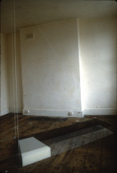 A long, cubic rectangle held by suspension wires on a weathered wooden floor. A small, white box rests within the rectangle.