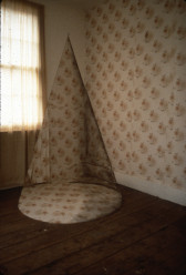 A large cone-shaped object in a room with wooden floors and a window covered with a sheer curtain. A light, floral print decorates one wallpapered wall; the same print covers the cone.