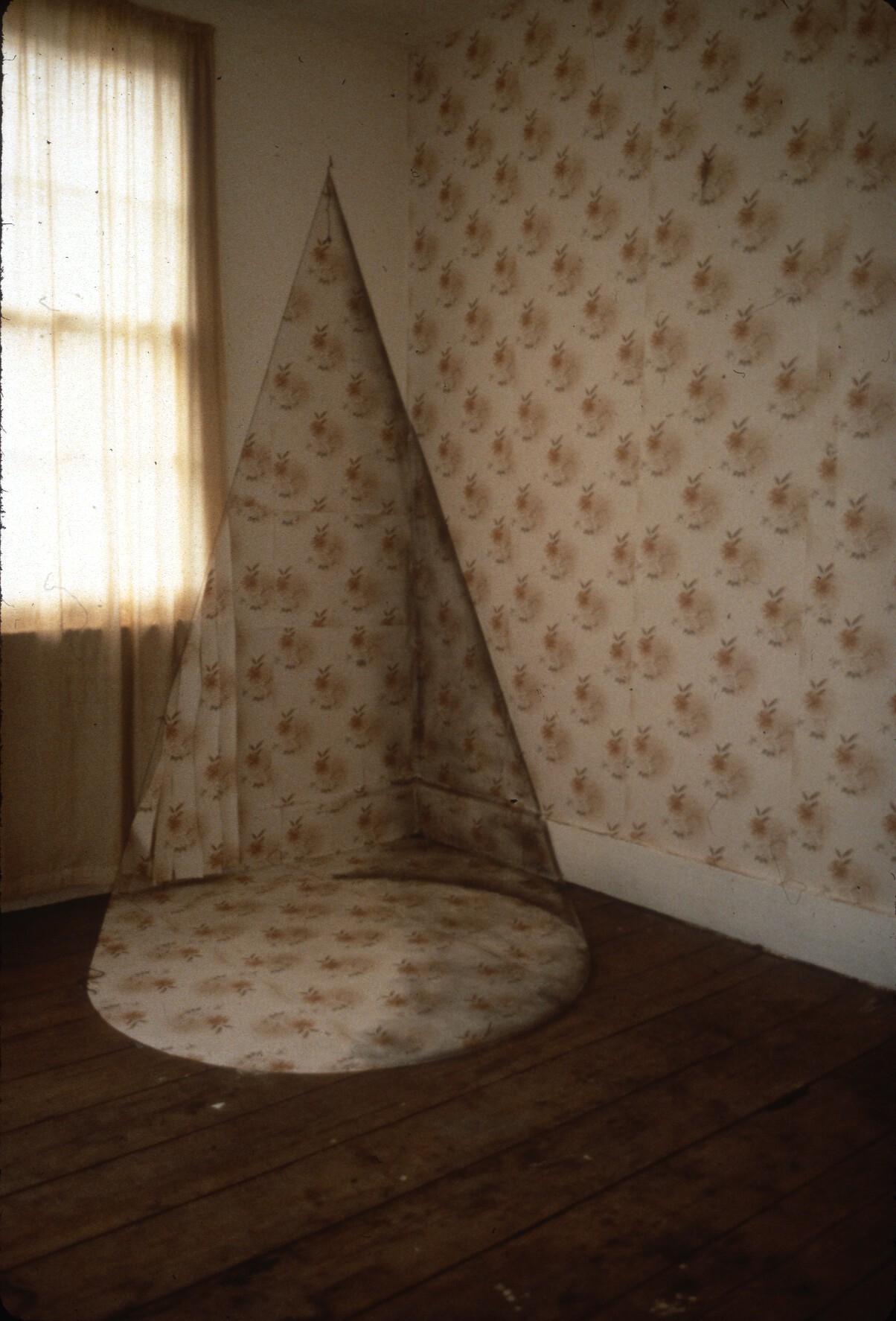 A large cone-shaped object in a room with wooden floors and a window covered with a sheer curtain. A light, floral print decorates one wallpapered wall; the same print covers the cone.