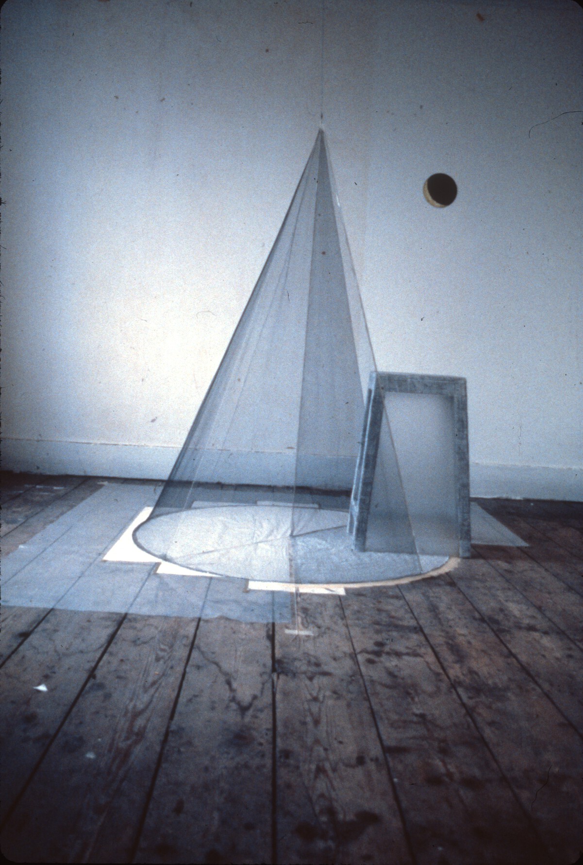 A transparent cone-shaped structure set in a bare room with wooden floors. There is a small hole in the wall just behind the structure.