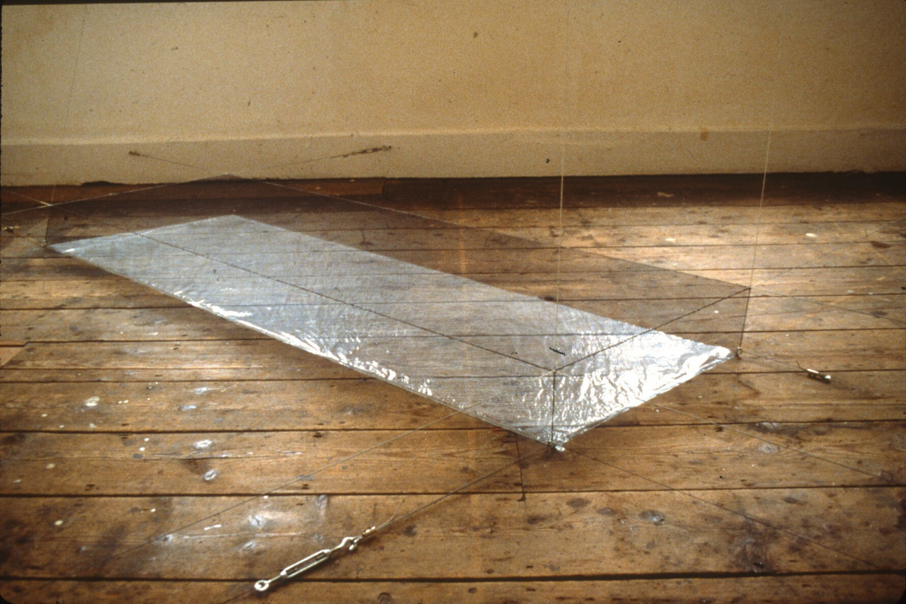 A long, cubic rectangle made from suspension wires with a silver, reflective base on a weathered wooden floor.