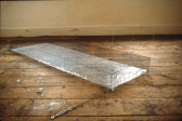 A long, cubic rectangle made from suspension wires with a silver, reflective base on a weathered wooden floor.