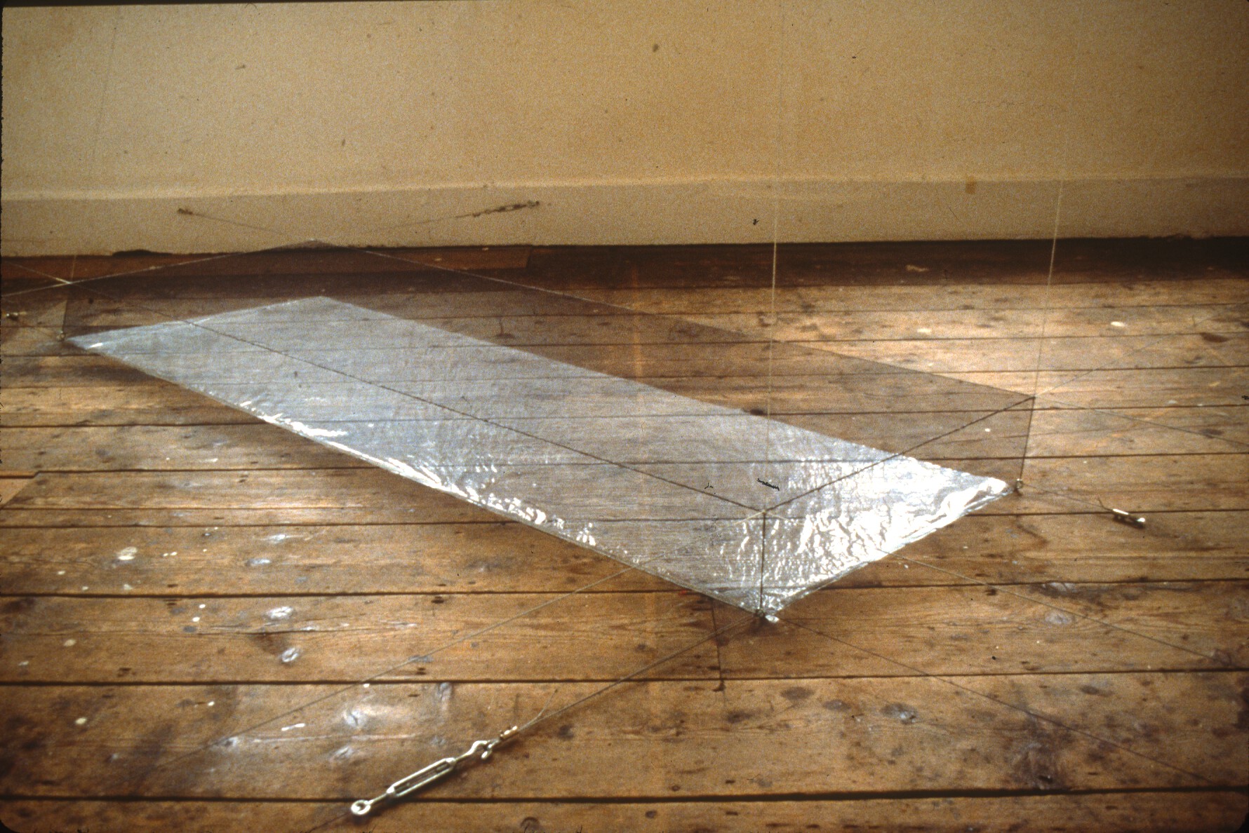 A long, cubic rectangle made from suspension wires with a silver, reflective base on a weathered wooden floor.