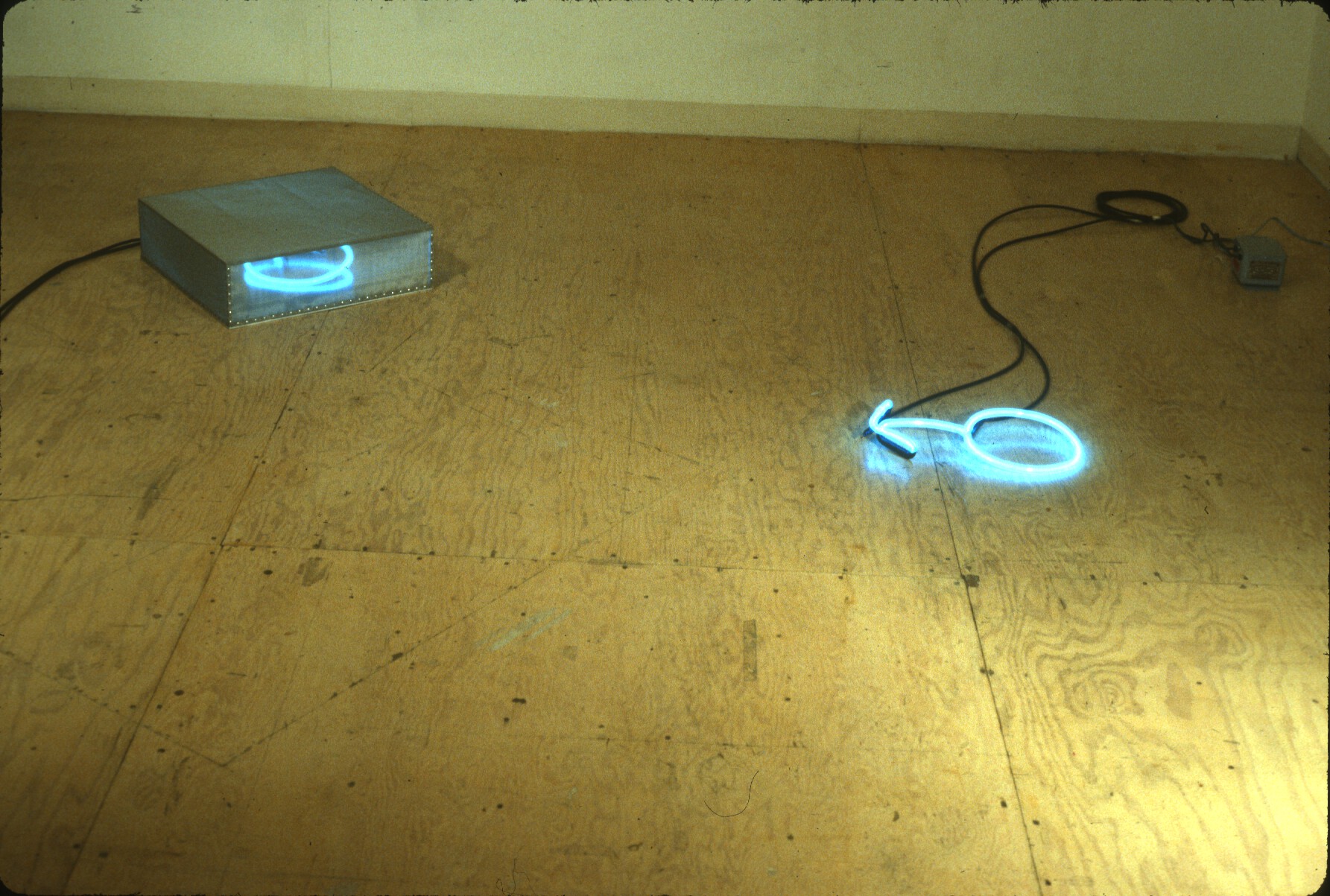 Two neon lights on a wooden floor. One light is coiled within a silver box, while the other rests directly on the floor, attached to a black cord.