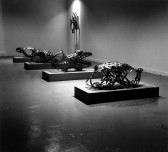 A black and white photograph of four sculptures resting on slightly elevated rectangular plinths, and a fifth sculpture mounted on the wall. The sculptures are constructed of metal molded into headless and limbless humanoid body shapes.
