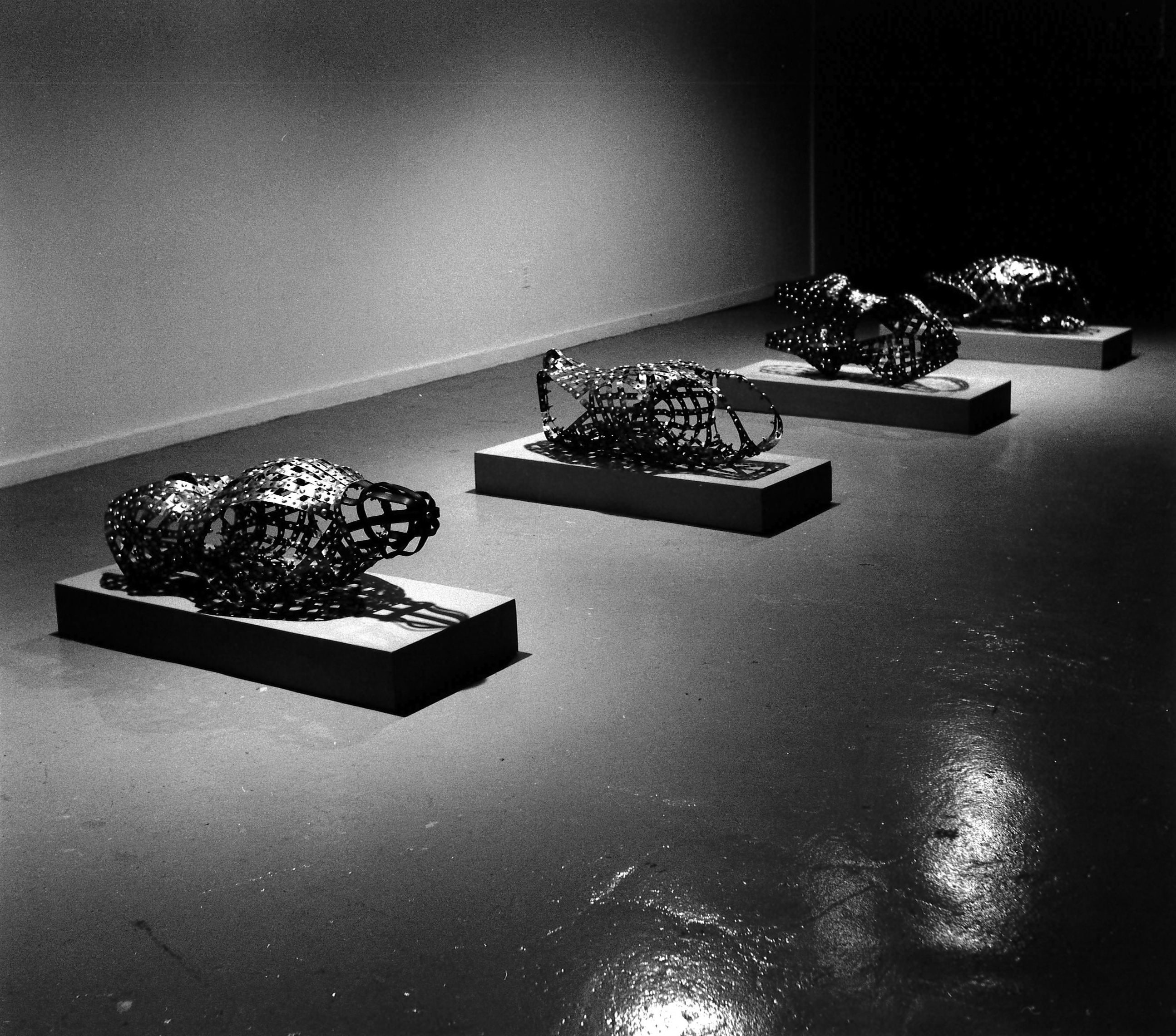 A black and white photograph of four sculptures resting on slightly elevated rectangular plinths. The sculptures are constructed of metal molded into headless and limbless humanoid body shapes.