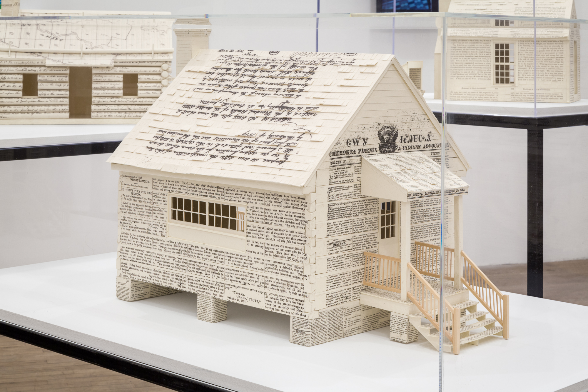 A miniature home crafted out of printed and handwritten material is displayed on a vitrine. At the front of the house above the doorway reads, 