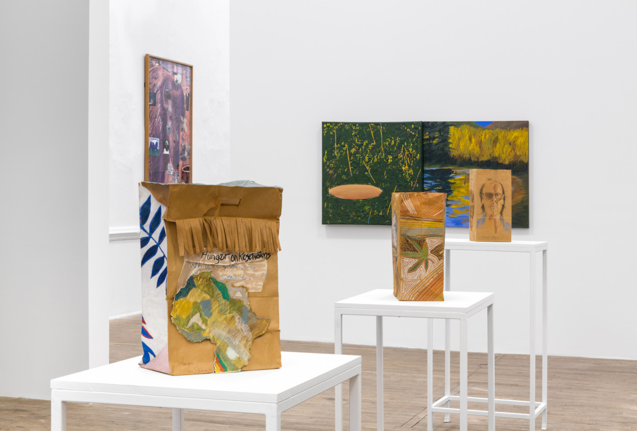 Three paper bags, decorated in collage and drawing, sit on pedestals of varying heights. An abstract lanscape painting hangs on a wall behind them. To the left, a purple painting of an buffalo hangs on another wall.