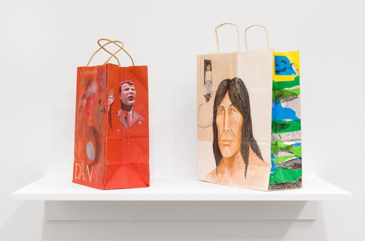 Two paper bags with handles sit on a white shelf. The left bag is red and decorated with two illustrated portraits, one of which is a Native American man. The bag to the right has a larger portrait of a Native American man next to illustrative fish on a colorful background.