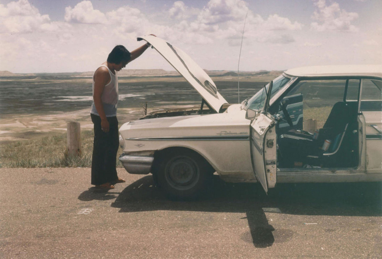 A man lifts up the hood of a white car on the side of the road amidst a sprawling open landscape. The front doors on both sides of the vehicle are open.