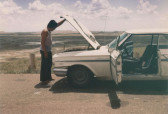 A man lifts up the hood of a white car on the side of the road amidst a sprawling open landscape. The front doors on both sides of the vehicle are open.