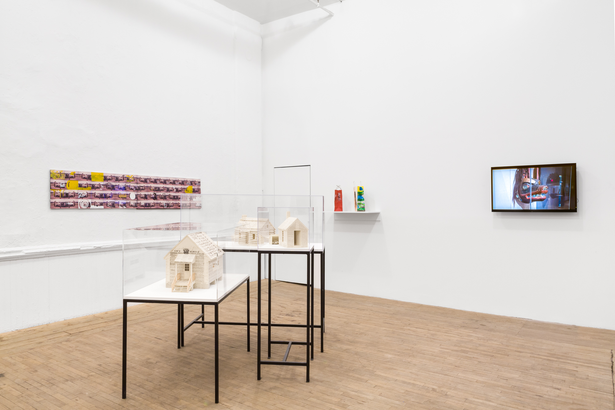 Miniature houses constructed of documents are displayed in the middle of the room on vitrines. A purple and yellow collage hangs on the back, left wall. The right wall displays colorful paper bags and a video monitor projecting an image of a robotic animal.