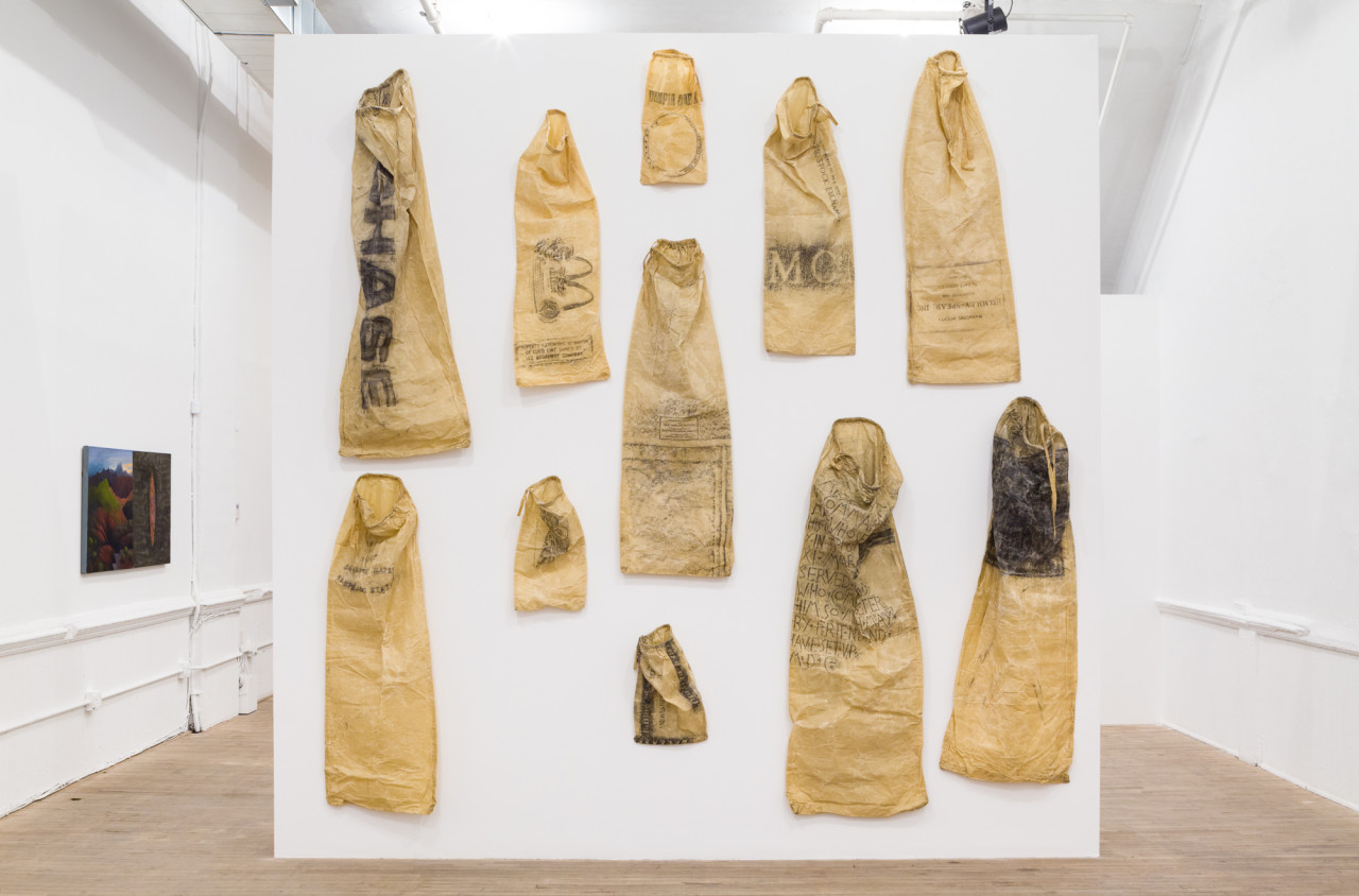 Eleven tan sacks of various sizes hang on a while wall. Each is illustrated with individual imagery and/or text. A long one, hanging at the top left, reads "CHASE" in block letters.  A painting is hung in the background on a wall.