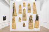Eleven tan sacks of various sizes hang on a while wall. Each is illustrated with individual imagery and/or text. A long one, hanging at the top left, reads 