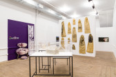 Miniature houses constructed of documents are displayed in vitrines in the middle of the room. Various photographs utilizing the wall and floor are displayed to the left on a purple section of wall. Tan, illustrated sacks of variable sizes hang on a white wall to the right.