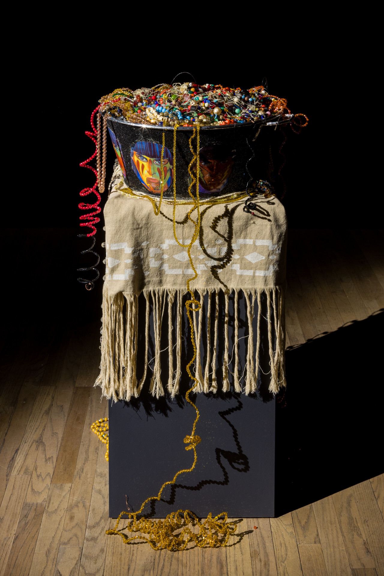 A large black bowl, painted with colorful faces, is filled to the brim with vibrant beaded jewelry. The bowl is placed on a tall black rectangular platform, covered with a beige woven blanket with long tassels and a white pattern along its border. Beaded jewelry from the bowl dangles down to the floor, intertwined and curled amongst themselves.