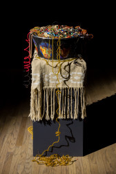 A large black bowl, painted with colorful faces, is filled to the brim with vibrant beaded jewelry. The bowl is placed on a tall black rectangular platform, covered with a beige woven blanket with long tassels and a white pattern along its border. Beaded jewelry from the bowl dangles down to the floor, intertwined and curled amongst themselves.