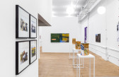 Four framed photogrpahs in a grid hang on an angled wall to the left. Three decorated paper bags sit on white pedestals. An abstract landscape painting hangs on the wall in the background.