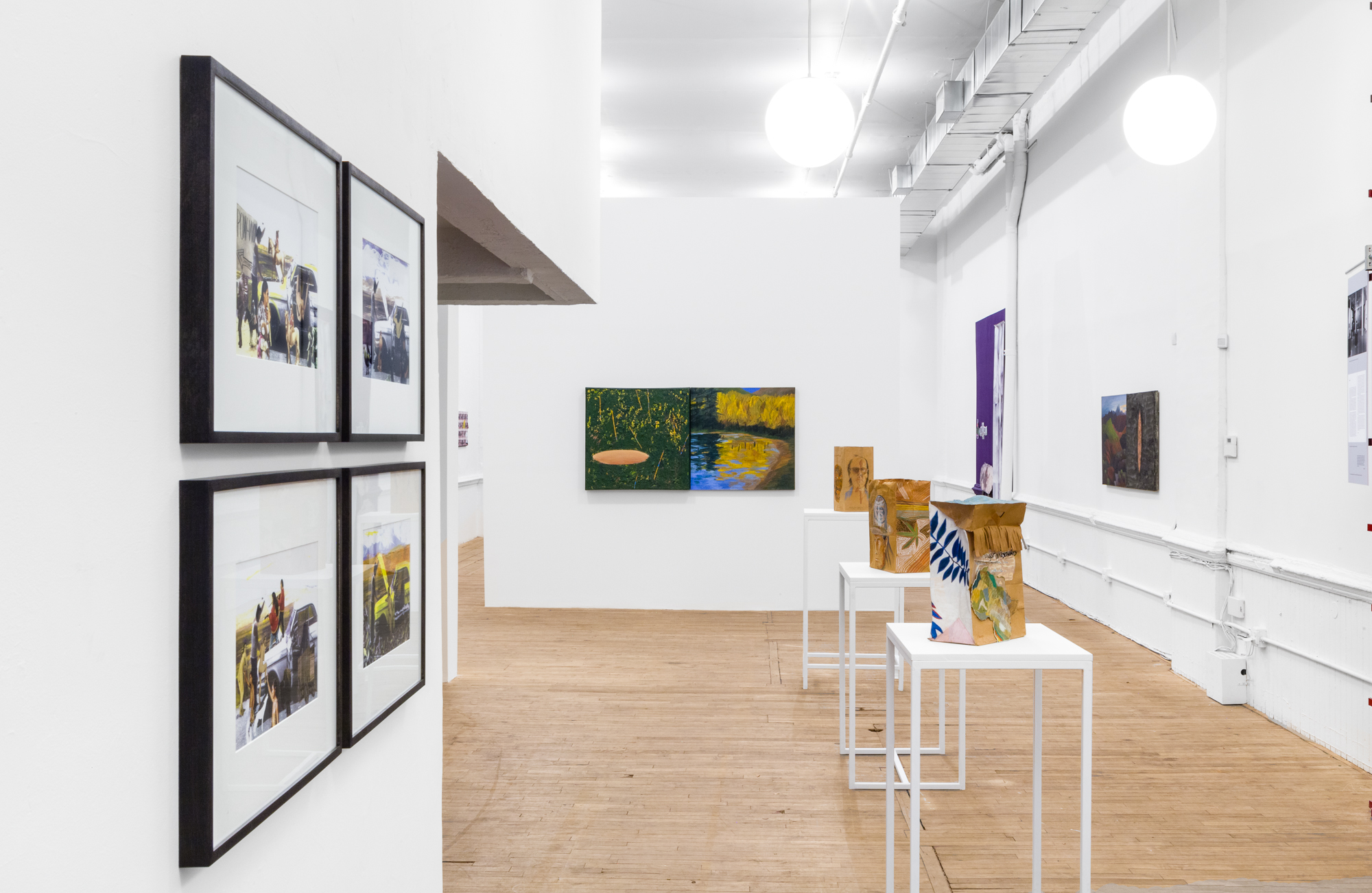 Four framed photogrpahs in a grid hang on an angled wall to the left. Three decorated paper bags sit on white pedestals. An abstract landscape painting hangs on the wall in the background.
