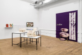 An angled view of two adjacent walls, in between sit vitrines with minature houses constructed from documents. From left to right on the wall: colorful paper bags, a monitor with a video still, a purple section of wall with various photographic objects.