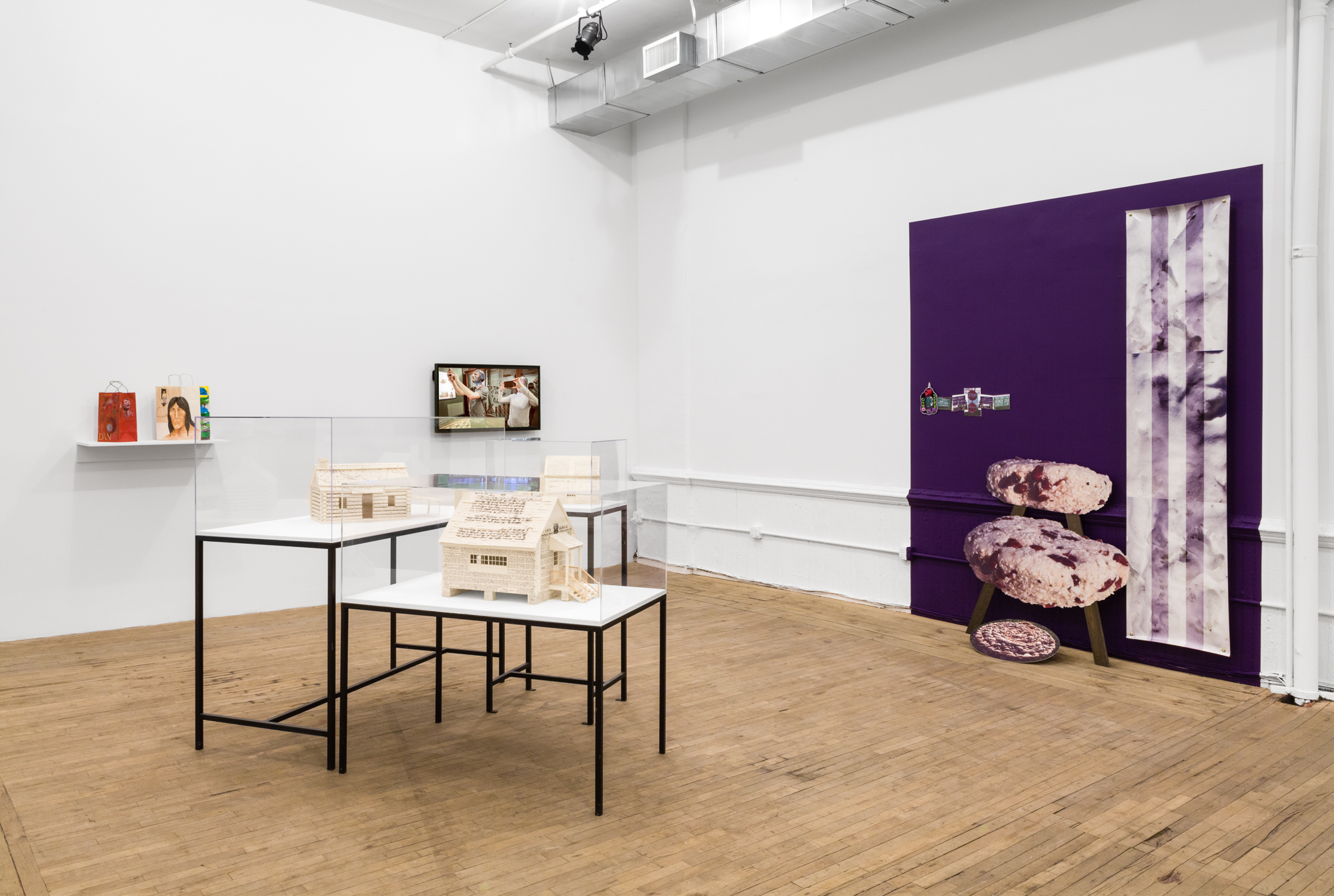 An angled view of two adjacent walls, in between sit vitrines with minature houses constructed from documents. From left to right on the wall: colorful paper bags, a monitor with a video still, a purple section of wall with various photographic objects.