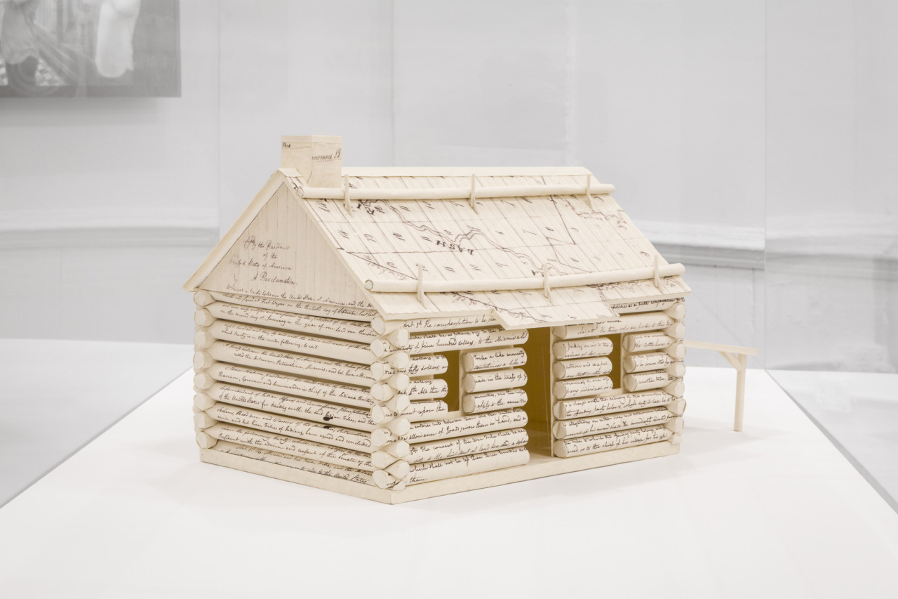A miniature log cabin constructed of handwritten documents is displayed in a vitrine. The roof on the left side reads, "By the President of the United States of America, A Proclimation."