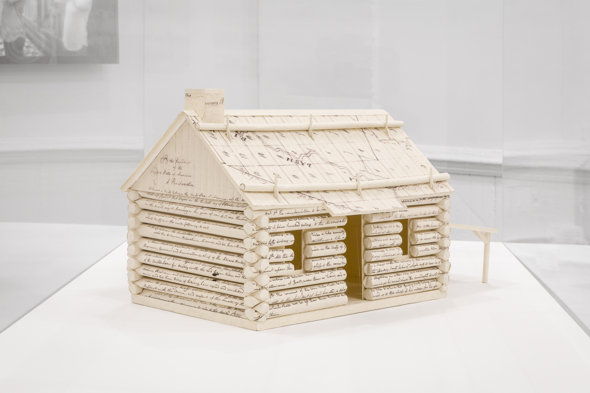 A miniature log cabin constructed of handwritten documents is displayed in a vitrine. The roof on the left side reads, 