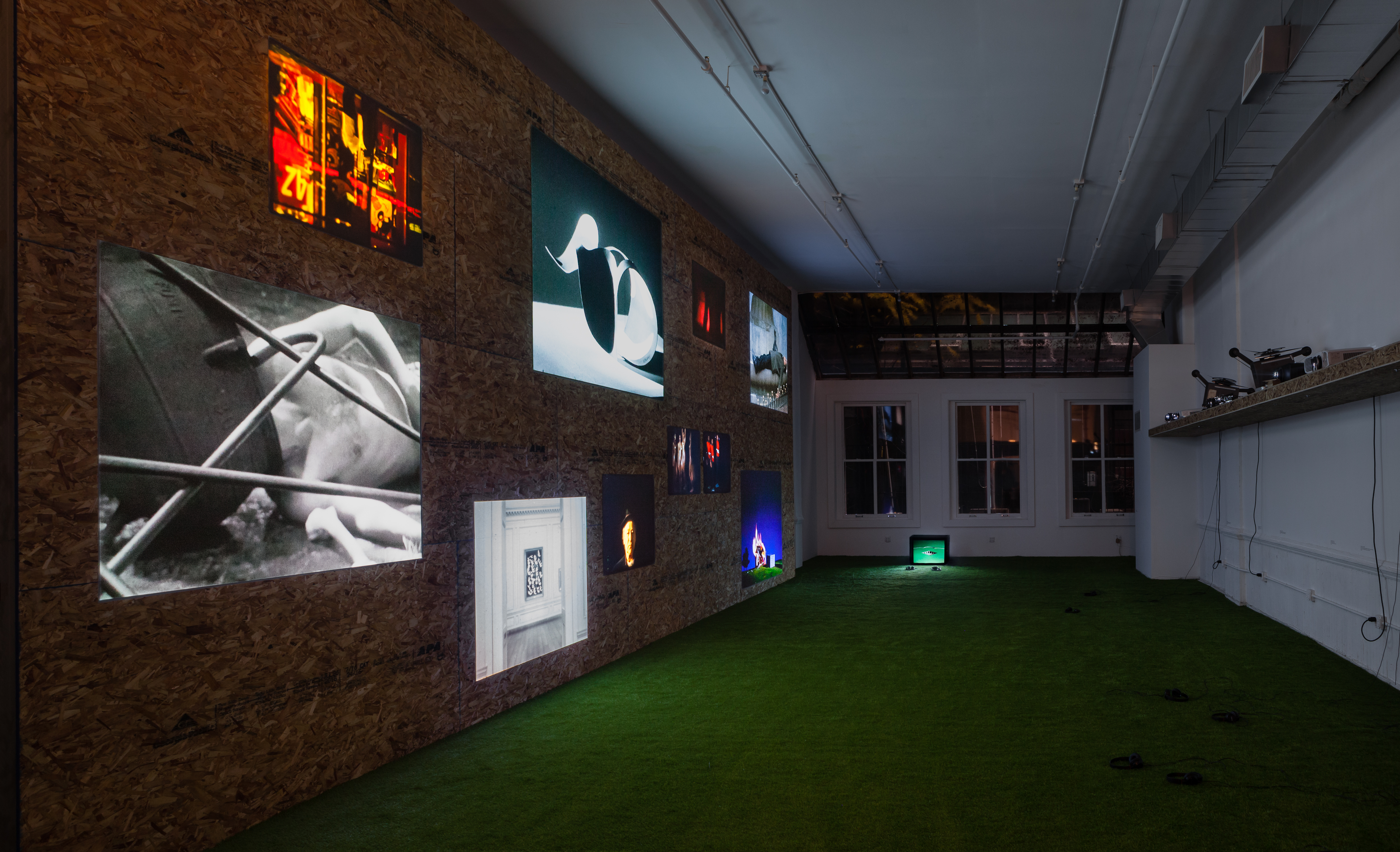 A dimly lit room with green turf-like carpet and exposed wooden walls. On the left, 9 images of different sizes are mounting, and luminous. On the right, projectors and movie cameras are placed on a horizontal shelf. On the far end of the floor towards the wall is a small square tv monitor with headphones placed in front of it. Behind the monitor are three medium-sized white windows.