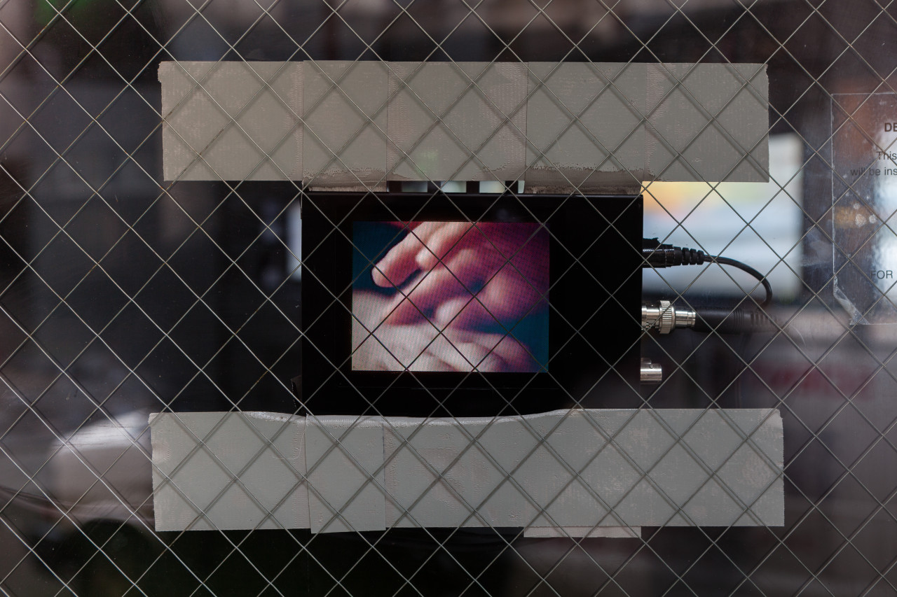 A small device with a screen and cables is stuck to a closed door with a glass window and safety guard. The screen displays a blurred close up image of two hands grazing one another.
