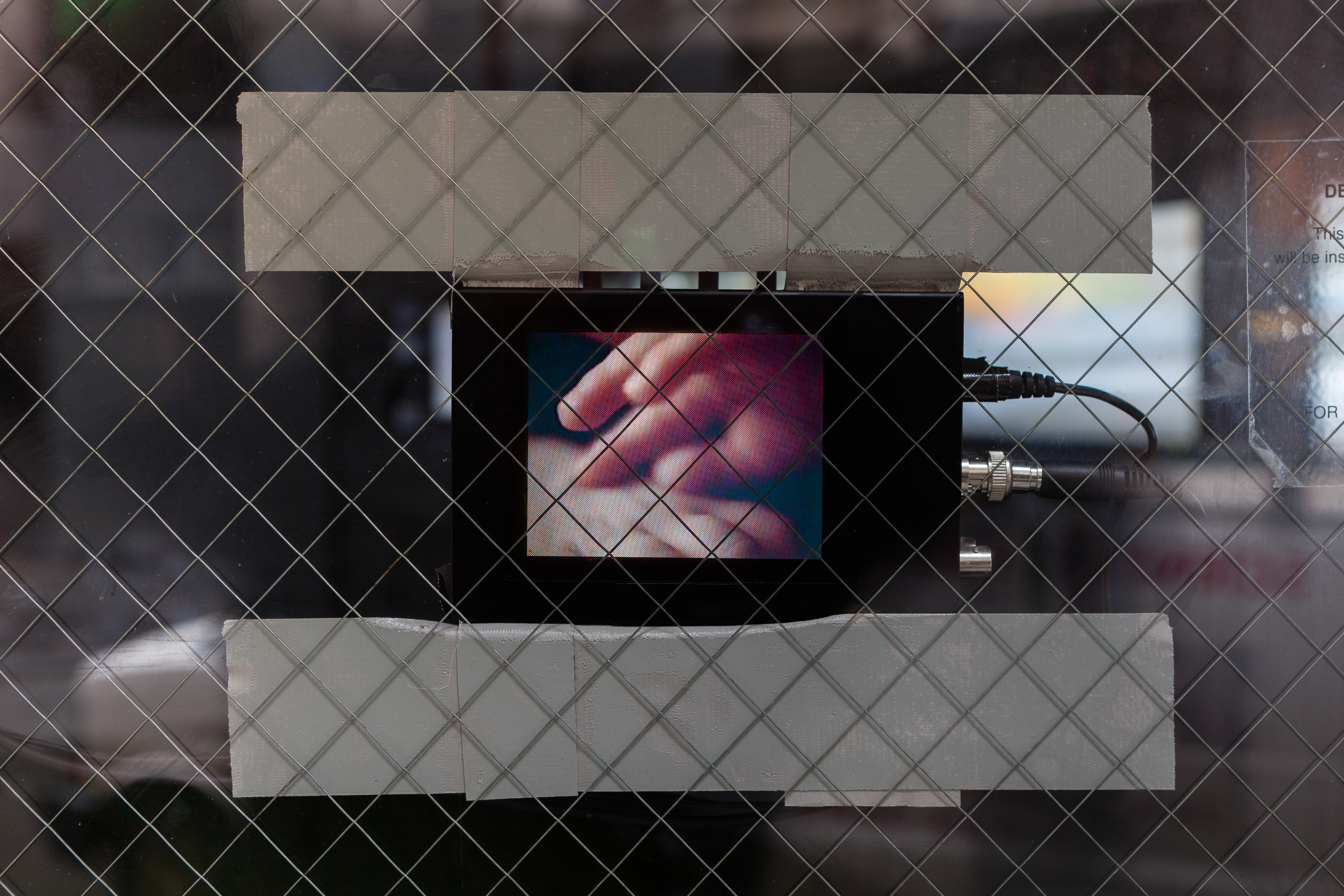 A small device with a screen and cables is stuck to a closed door with a glass window and safety guard. The screen displays a blurred close up image of two hands grazing one another.