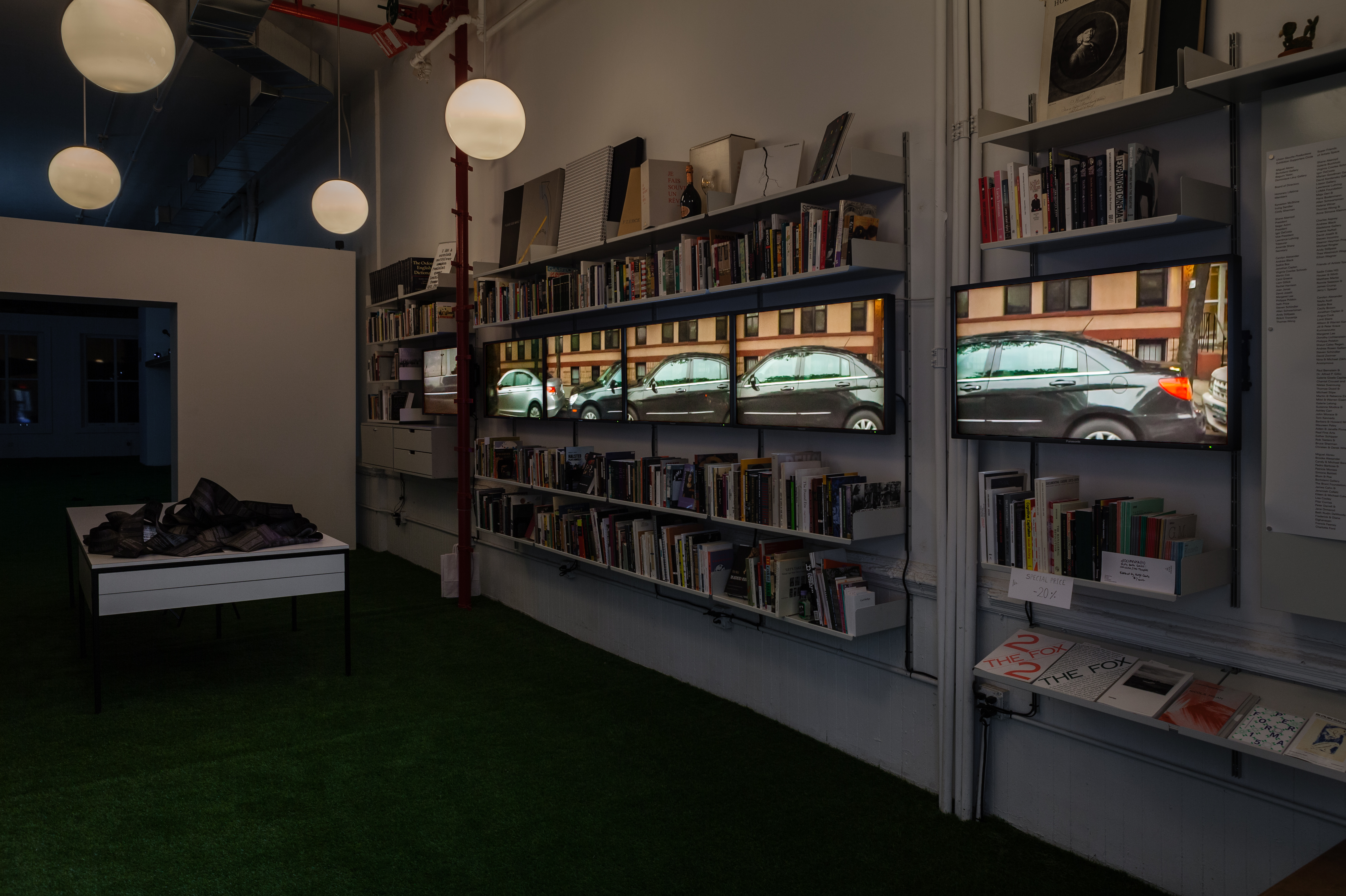 A diagonal view of six monitors in a row are lined up in the middle of a floating bookshelf, displaying stills from the film. On the left corner of the image, a desk holds long slips of black paper with white text on it, curled and intertwined, resembling film strips.