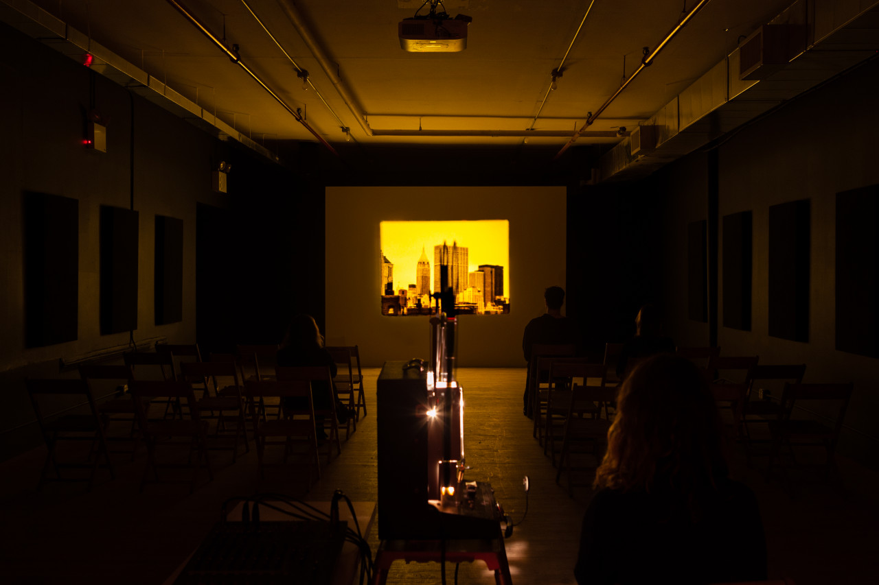 A dimly lit room where two people are seated in chairs on the right, looking at a projection. The projection displays a yellow-hued image of a city skyline.