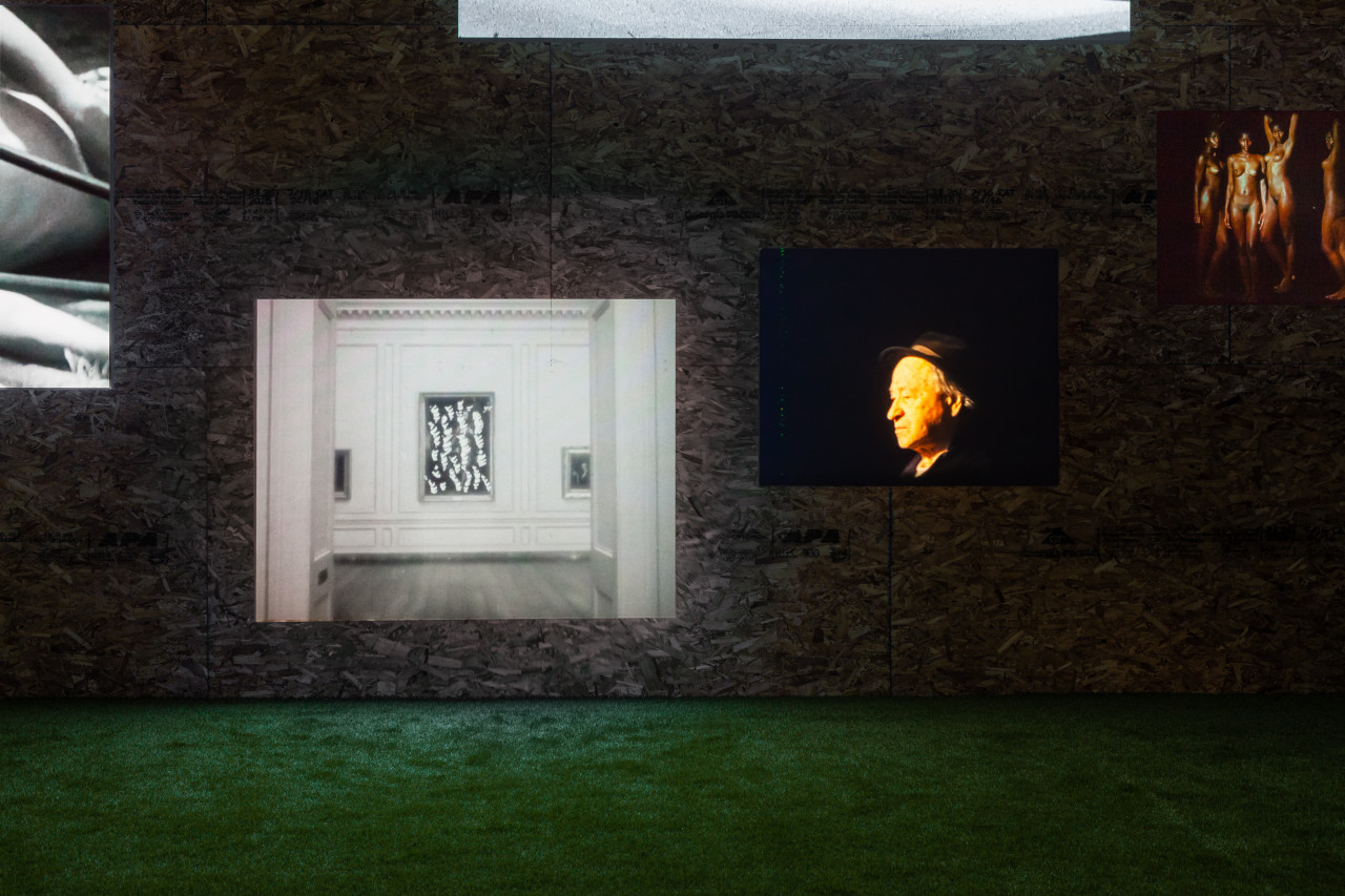 Five projections playing on a wall at different heights. Two are completely visible and three are partially visible. In the center of the image is a black and white projection of a gallery, with a painting placed in between the entrance of a hallway. To its right is a projection of a Jonas Mekas facing left, his face slightly golden in tone, wearing a fedora. A projection to its right partially shows four dark skinned people, nude and posing.