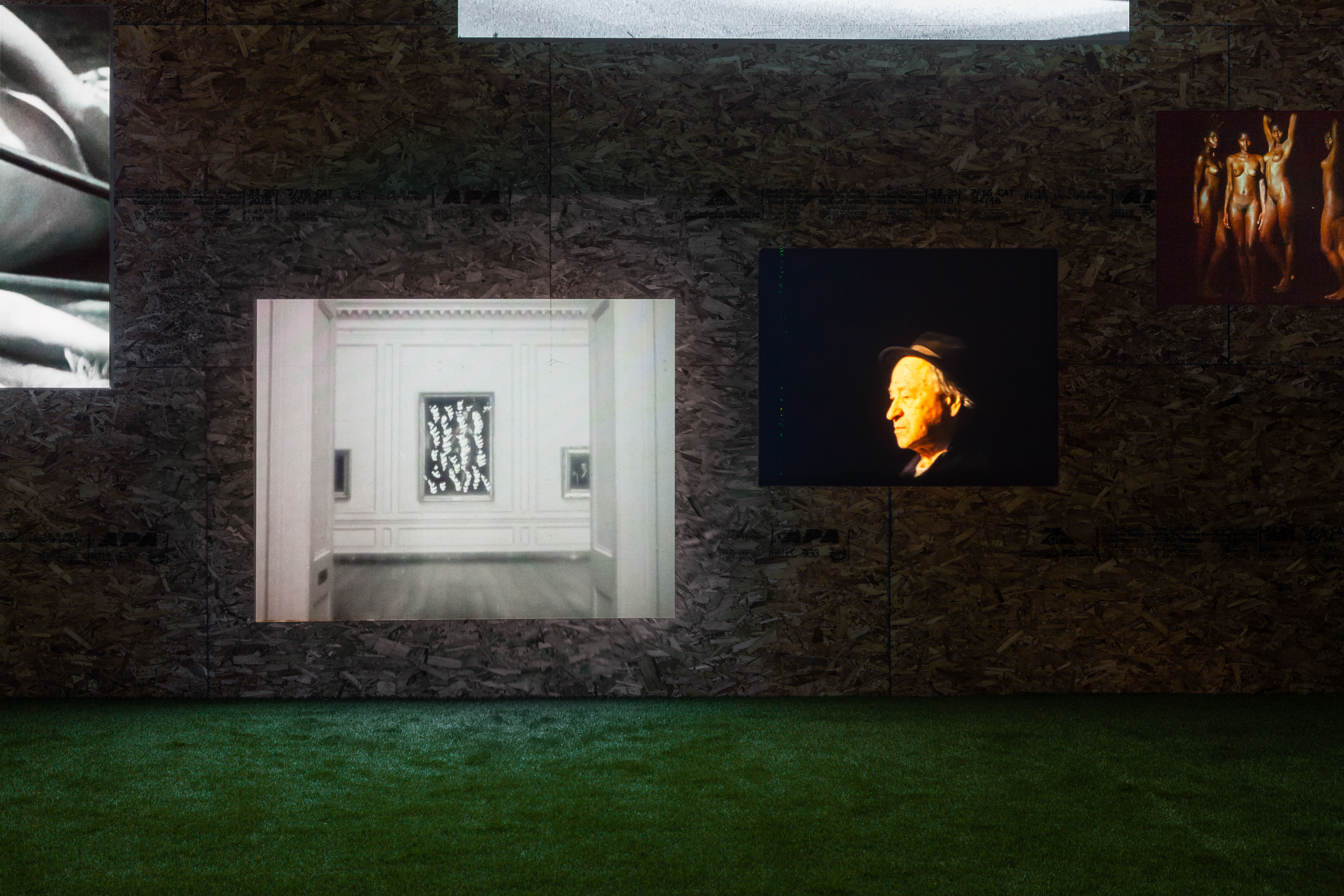 Five projections playing on a wall at different heights. Two are completely visible and three are partially visible. In the center of the image is a black and white projection of a gallery, with a painting placed in between the entrance of a hallway. To its right is a projection of a Jonas Mekas facing left, his face slightly golden in tone, wearing a fedora. A projection to its right partially shows four dark skinned people, nude and posing.