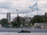 A man lies on the street in a horizontal position, centered within the frame. Behind him is a playground, with an Argentinian flag waving in the background.