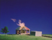 A still from Totloop, set during the twilight, featuring an empty model house being lit on fire, with a small tree visible to its left.