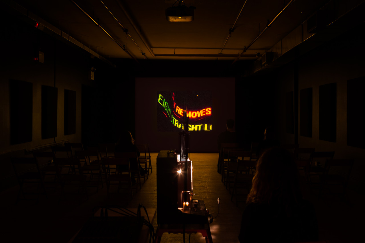 A dimly lit room with a projection facing the front. The projection displays neon words that are slightly distorted, the legible text reads "Removes.....eight straight...."