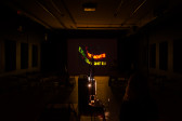 A dimly lit room with a projection facing the front. The projection displays neon words that are slightly distorted, the legible text reads 
