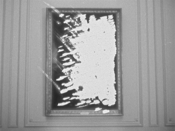 A black and white video still of a painting hanging on a decorative wall. The image in the painting has been burned and is illegibly white.