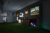 An angled view of scattered video projection stills at various heights and sizes on a wooden wall. The wooden wall is displayed in a room with white walls and faux-grass flooring.